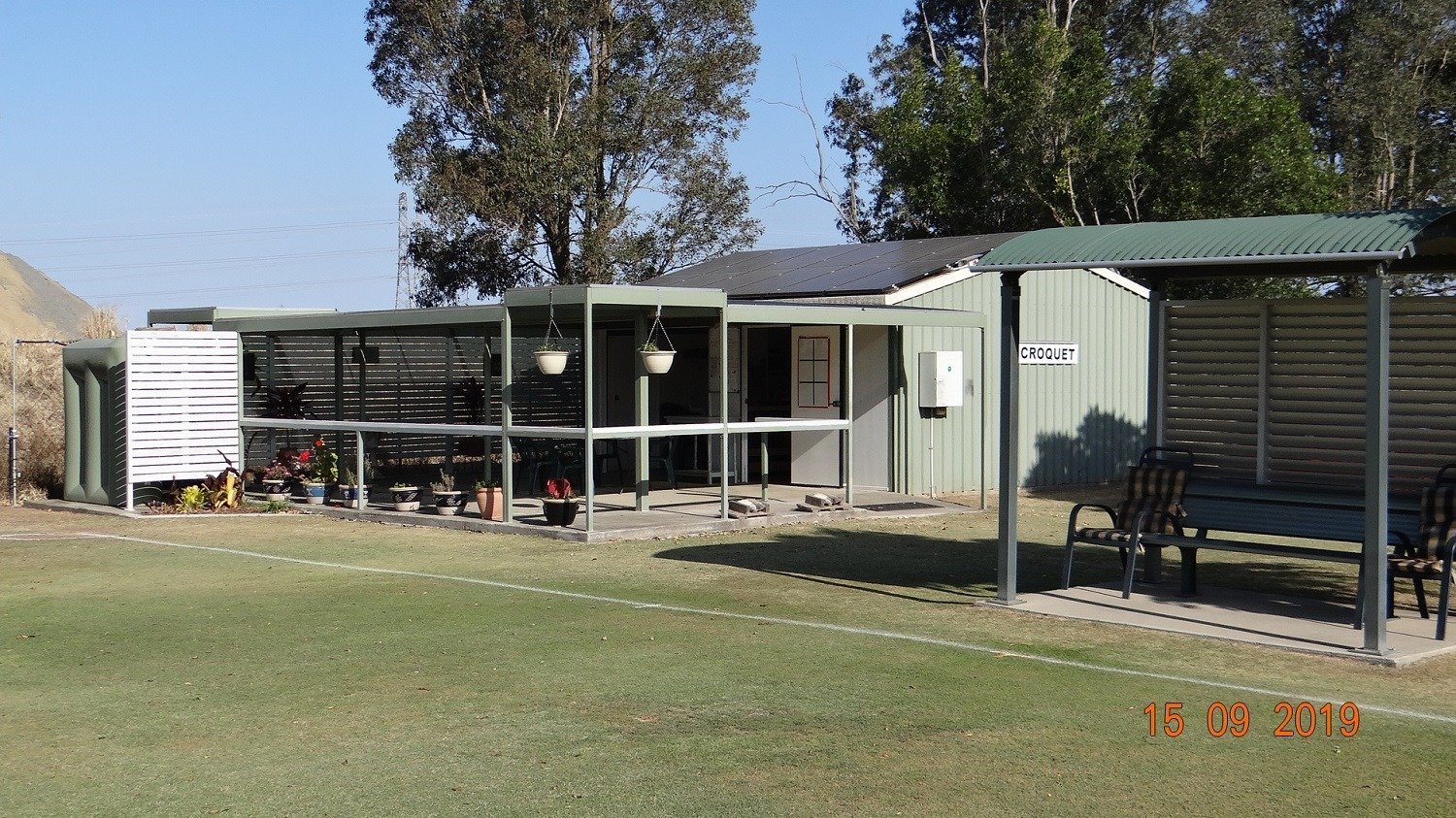 Croquet club lawn with clubhouse building, covered seating area, and tall eucalyptus trees in background