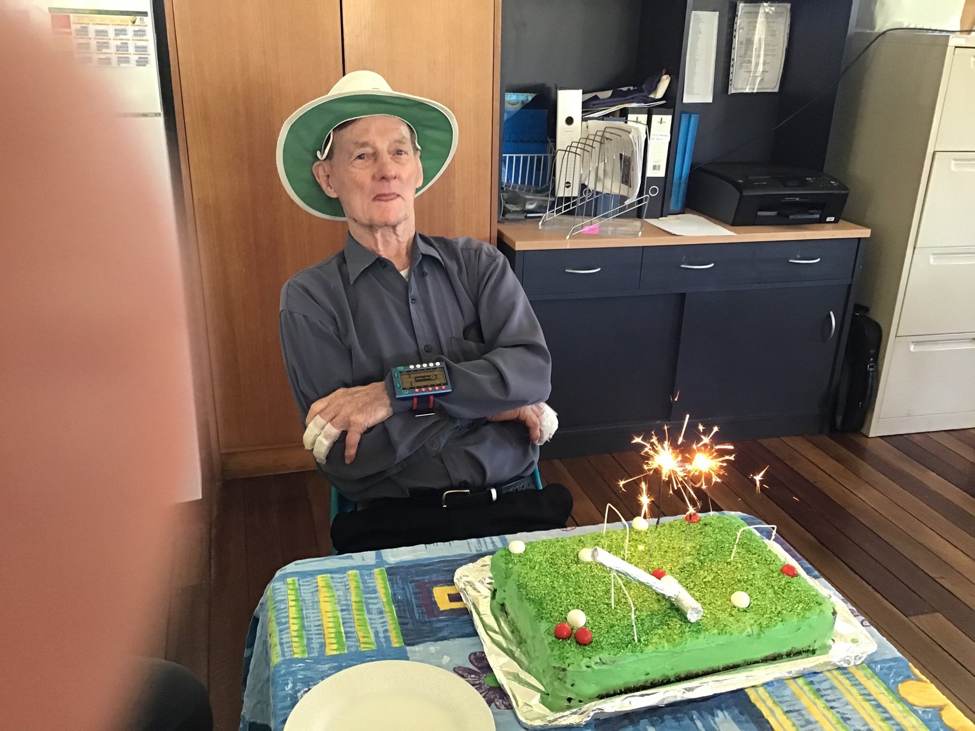 Man in green hat seated at table with croquet-themed cake, sparkler, and scoreboard in club hall