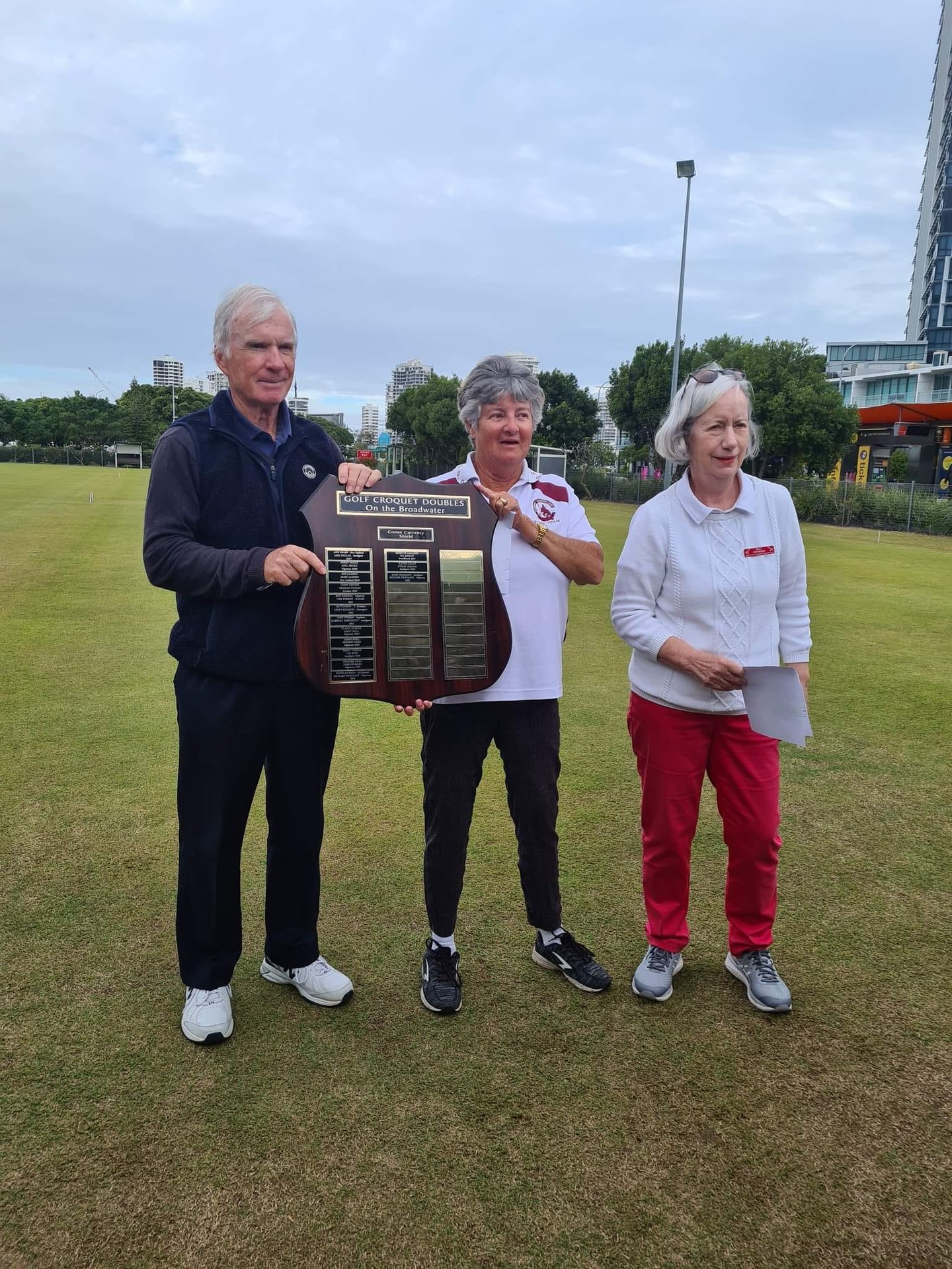 Three players holding a trophy on a croquet lawn with city buildings in background