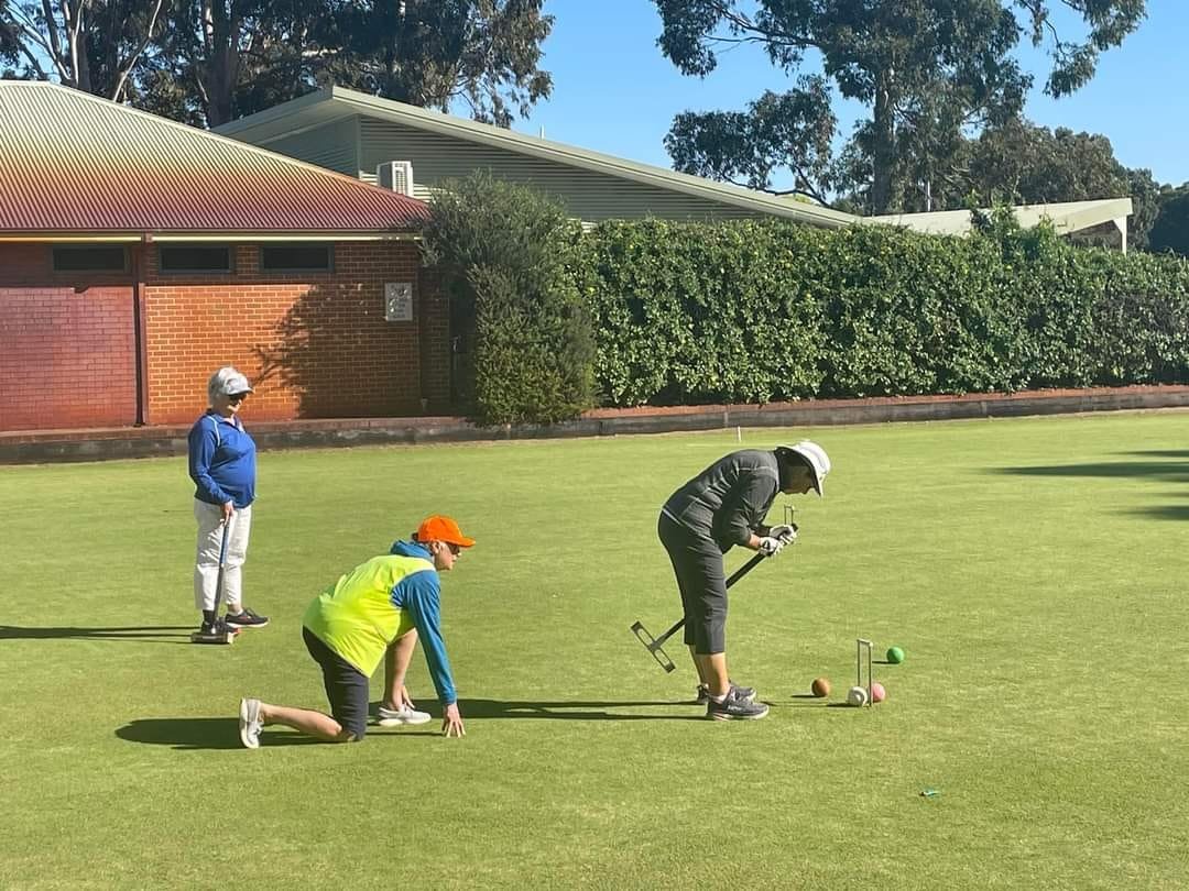 Players on club lawn, one preparing to strike ball through hoops with mallet while others watch