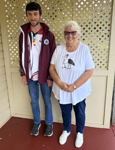 A young man wearing a medal stands beside an older woman in a t-shirt featuring a bird holding a croquet mallet