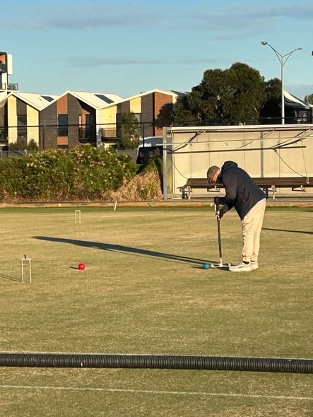 A croquet player bends over to strike a blue ball with a mallet on a manicured lawn with wickets