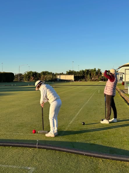 A croquet player in white prepares to strike a red ball with a mallet while another player watches on a sunny lawn