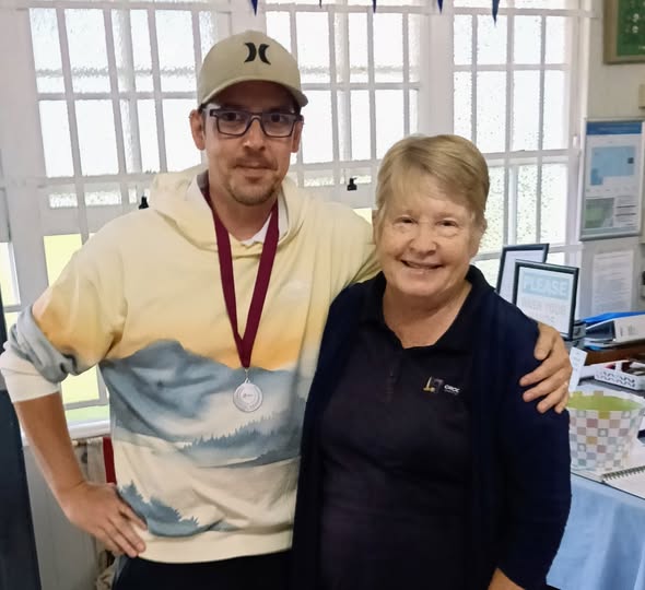 A man wearing a silver medal and a cap poses with a smiling woman in a blue shirt with a croquet mallet logo