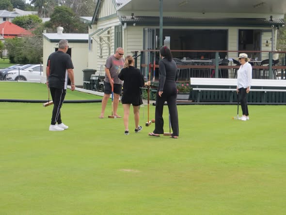 Five people holding croquet mallets stand on a green lawn in front of a clubhouse