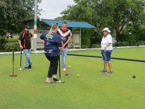 Four people stand on a croquet lawn holding mallets near red, yellow, and black balls and a wicket