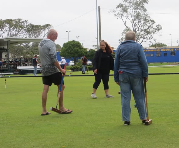 Three people stand on a croquet lawn holding mallets near a blue building labeled Wynnum Bowls Club