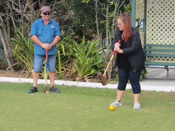 A woman swings a croquet mallet at a yellow ball on a lawn while a man in blue watches