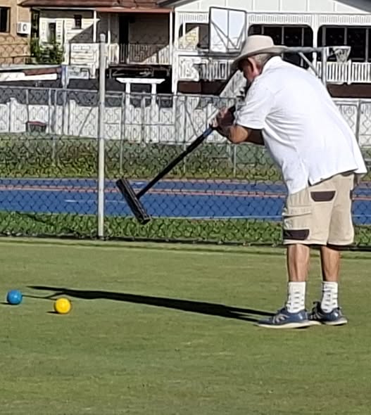 A man in a hat and white shirt swings a croquet mallet on a green lawn near blue and yellow balls