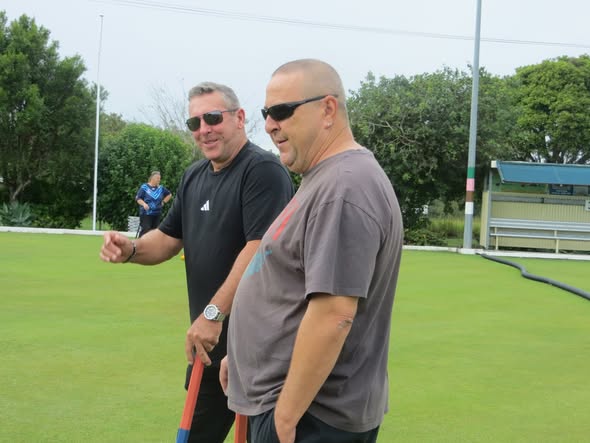 Two men wearing sunglasses stand on a croquet lawn, one holding a mallet and gesturing