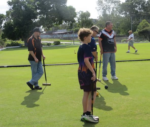 A boy holds a croquet mallet on a green lawn while other players stand nearby with mallets and balls