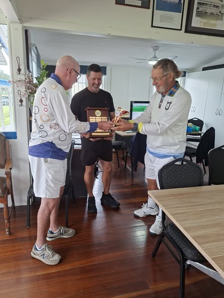 Three men, two in white croquet attire, stand in a clubhouse exchanging a wooden plaque and a gold trophy