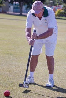 A croquet player in white attire holds a mallet on a grass court near a red ball