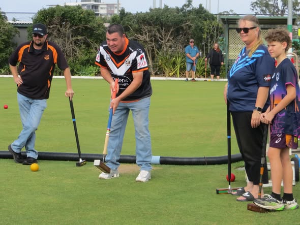 A man in a Wests Tigers jersey swings a croquet mallet on a green lawn near a boundary hose while others watch