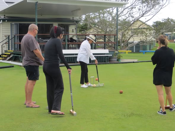 Four people on a croquet lawn watching a woman in a white hat strike a ball with a mallet