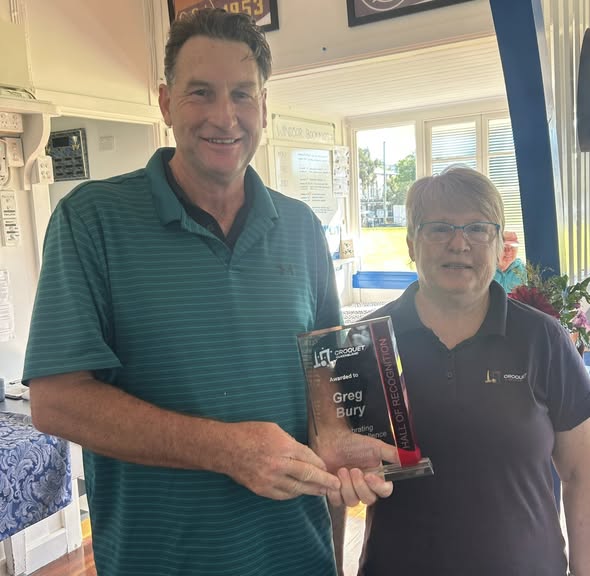 Greg Bury and a woman stand inside a clubhouse holding a Croquet Hall of Recognition award