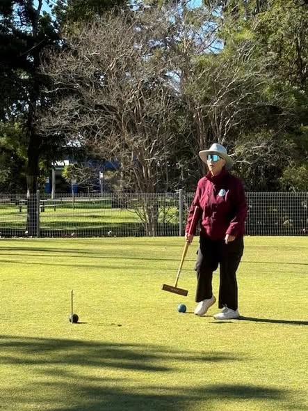 A woman in a maroon shirt and hat stands on a croquet lawn holding a mallet near a blue ball and a peg
