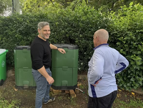 George Clooney smiles at a man in a white and blue croquet shirt standing near green compost bins
