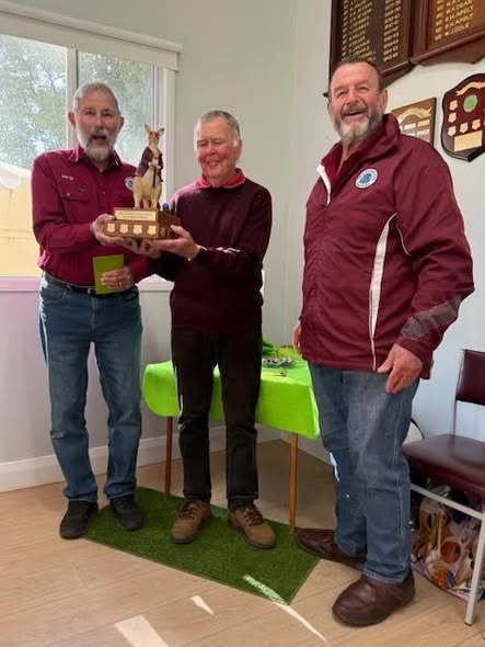 Three men stand indoors, two holding a croquet trophy topped with a mallet and ball, standing on a green mat