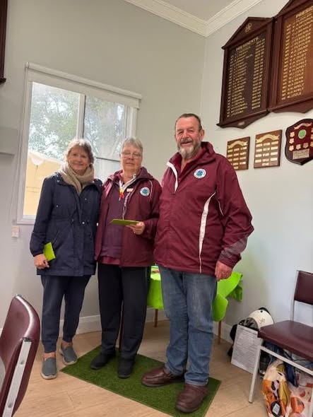 Three croquet club members stand on an indoor green mat holding certificates near wooden honor boards