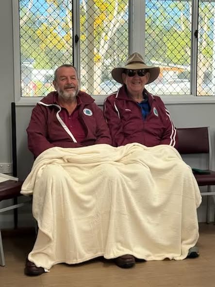 Two men in maroon croquet club jackets sit on a bench sharing a cream blanket