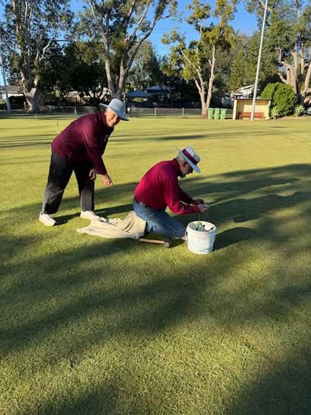 Two people on a croquet lawn, one kneeling near a bucket and mallet while the other stands and points