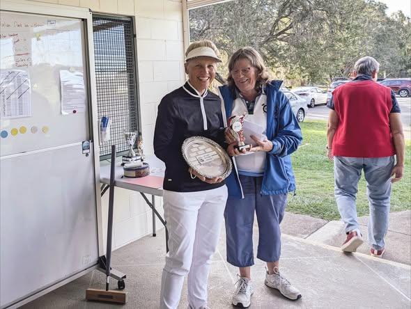 Two women in croquet attire hold trophies and a silver plate beside a table with a mallet and awards