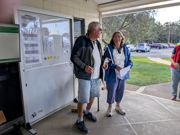 A man holding a croquet mallet and a woman holding an award stand by a whiteboard with tournament sheets
