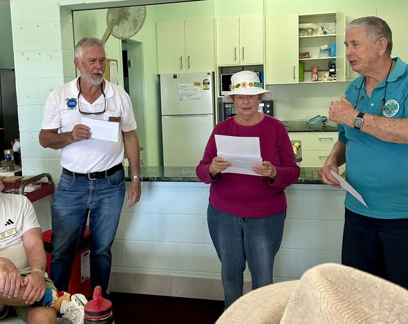 Three croquet club members stand in a kitchen, holding papers, likely reviewing scores or rules during a club meeting