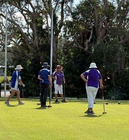 Four players in team uniforms play croquet on a sunny lawn, with mallets and balls visible near wickets