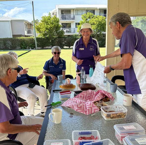 Croquet players in purple uniforms gather around a table with food and drinks at a club, with a croquet court visible in the background