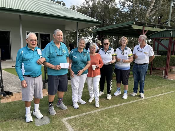 Seven people in matching teal shirts stand on a croquet lawn, holding a certificate and cups, with a clubhouse in the background