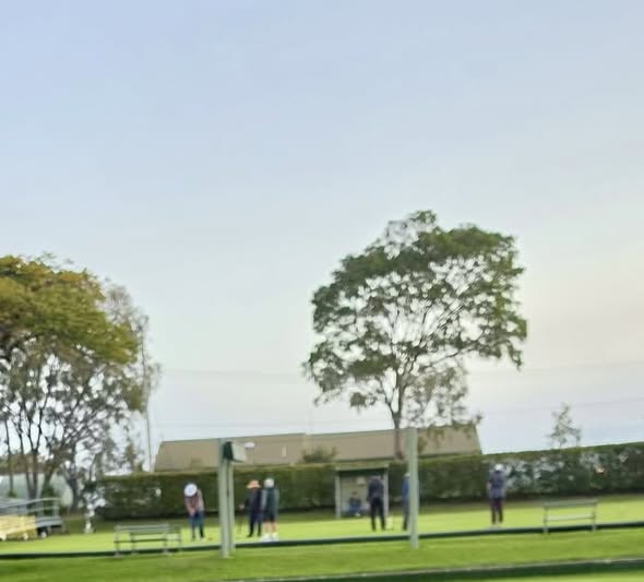 People play croquet on a grassy field with wickets and mallets, near a building and trees under a clear sky