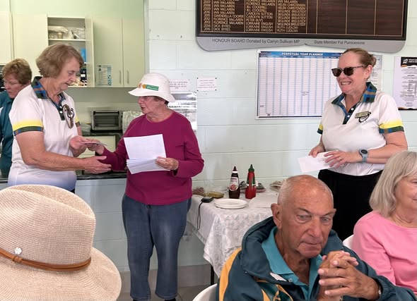 Women in croquet uniforms receive awards at a club event, with one holding a certificate and a scoreboard visible in the background