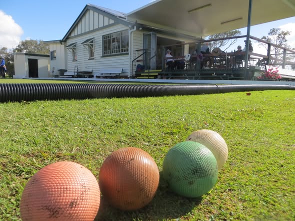 Four croquet balls—orange, green, and cream—rest on a grassy lawn near a black pipe, with a clubhouse and spectators in the background