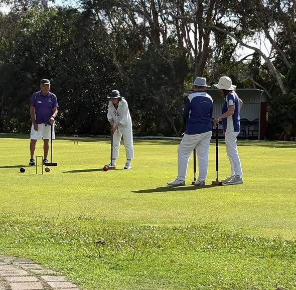 Four players in croquet attire play on a sunny lawn, with one in white preparing to strike a ball with a mallet near a wicket