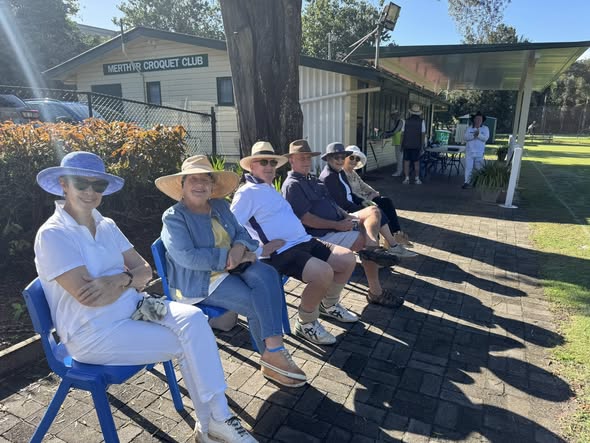 Spectators in sun hats sit on blue chairs at the Merthyr Croquet Club, watching a game under sunny skies