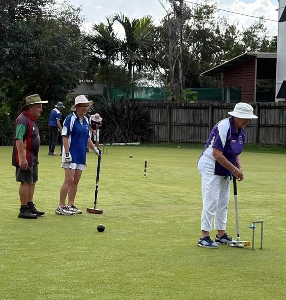 A woman in a purple shirt prepares to hit a croquet ball with a mallet on a grassy court, with other players watching