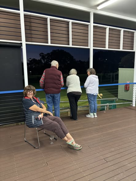 A woman sits on a chair on a deck, watching three people stand and observe a croquet game at night through a glass window