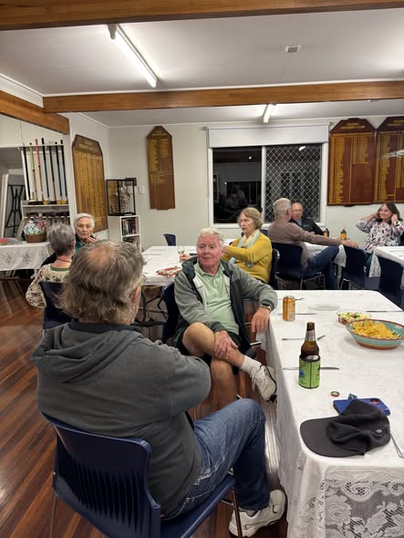 Adults socializing at a croquet club hall, seated at tables with drinks and food, surrounded by croquet equipment and trophy boards