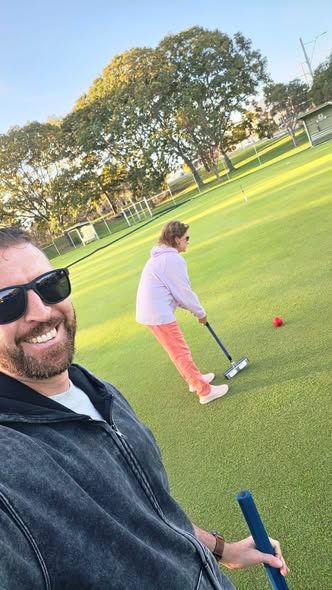 A man takes a selfie while playing croquet, with a woman in the background preparing to hit a red ball with her mallet on a sunny lawn