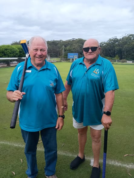 Two men in matching teal croquet shirts stand on a grassy croquet lawn, each holding a croquet mallet