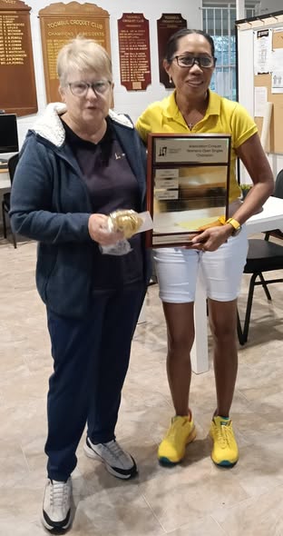 Two women pose indoors at the Toombul Croquet Club, one holding a framed certificate and the other a trophy, celebrating a croquet tournament win