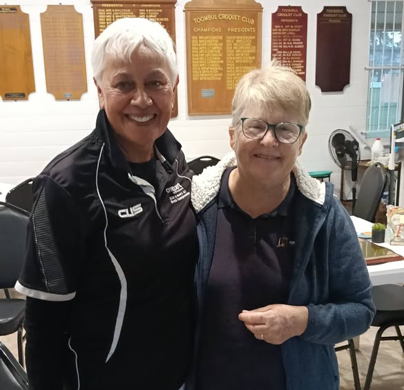 Two women smile at the camera inside the Toombul Croquet Club, with championship plaques visible on the wall behind them