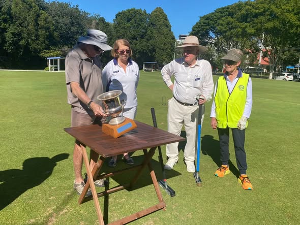 Four people in croquet attire gather around a trophy on a wooden table on a sunny lawn, with mallets nearby