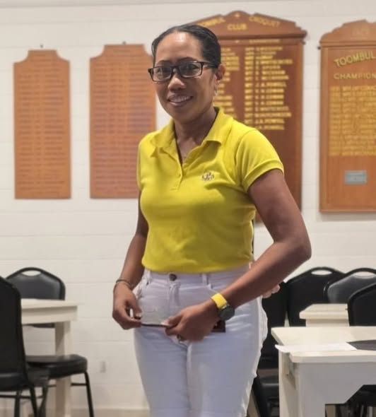 A woman in a yellow polo and white pants stands indoors at a croquet club, holding a mallet, with championship plaques on the wall behind her