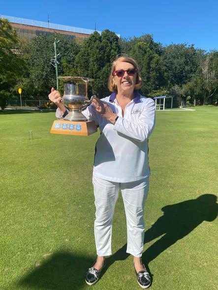A woman in white croquet attire holds a large silver trophy on a sunny croquet lawn, celebrating a victory