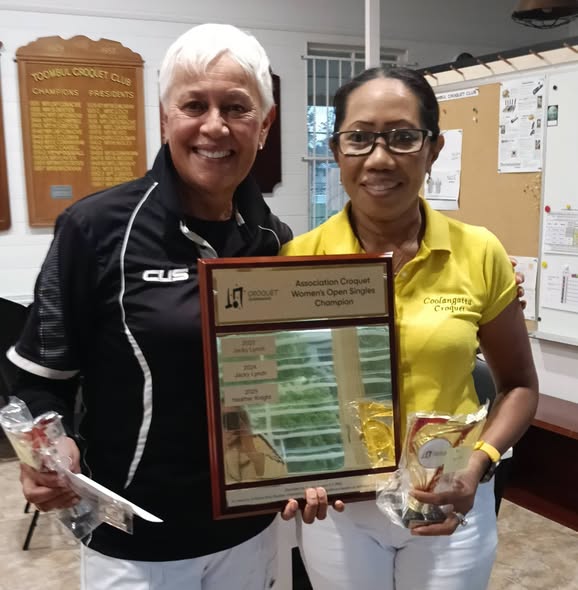 Two women pose with a framed award for the Association Croquet Women's Open Singles Champion, holding trophies in a club setting