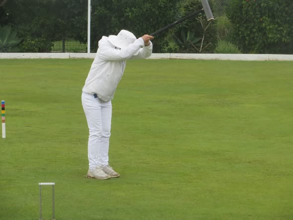 A person in white croquet attire swings a mallet at a ball on a green lawn, with a wicket and peg visible nearby