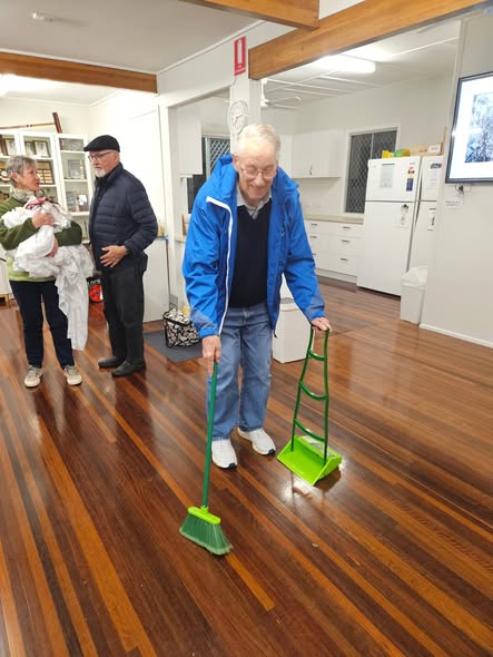 An older man in a blue jacket uses a green croquet mallet and ball indoors on a wooden floor, with others watching